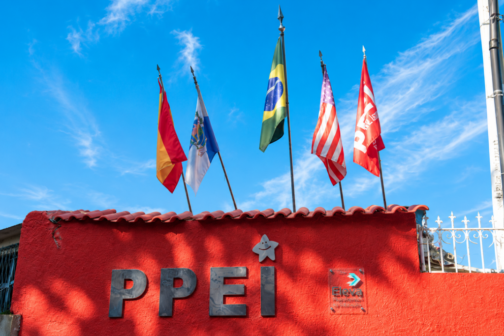 Fachada da PPEI Escola Bilíngue no Méier, com bandeiras e céu azul.
